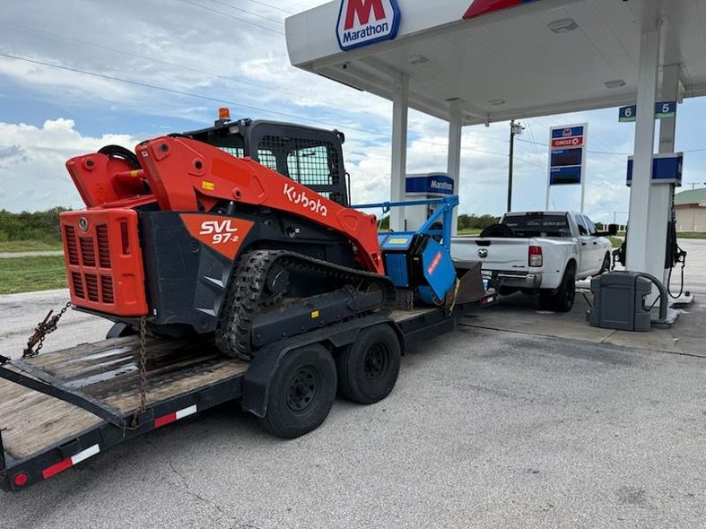 Orange skid steer on a trailer at a gas station pump, with a pickup truck nearby.