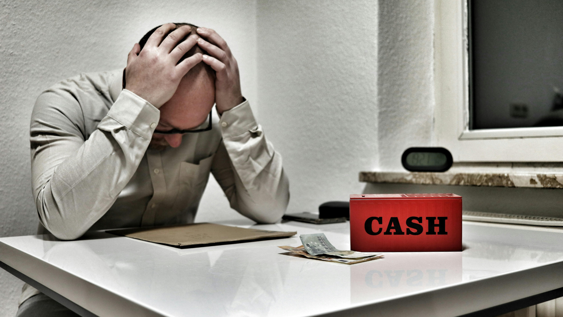 Stressed person at a desk with head in hands beside a red cash box and scattered papers