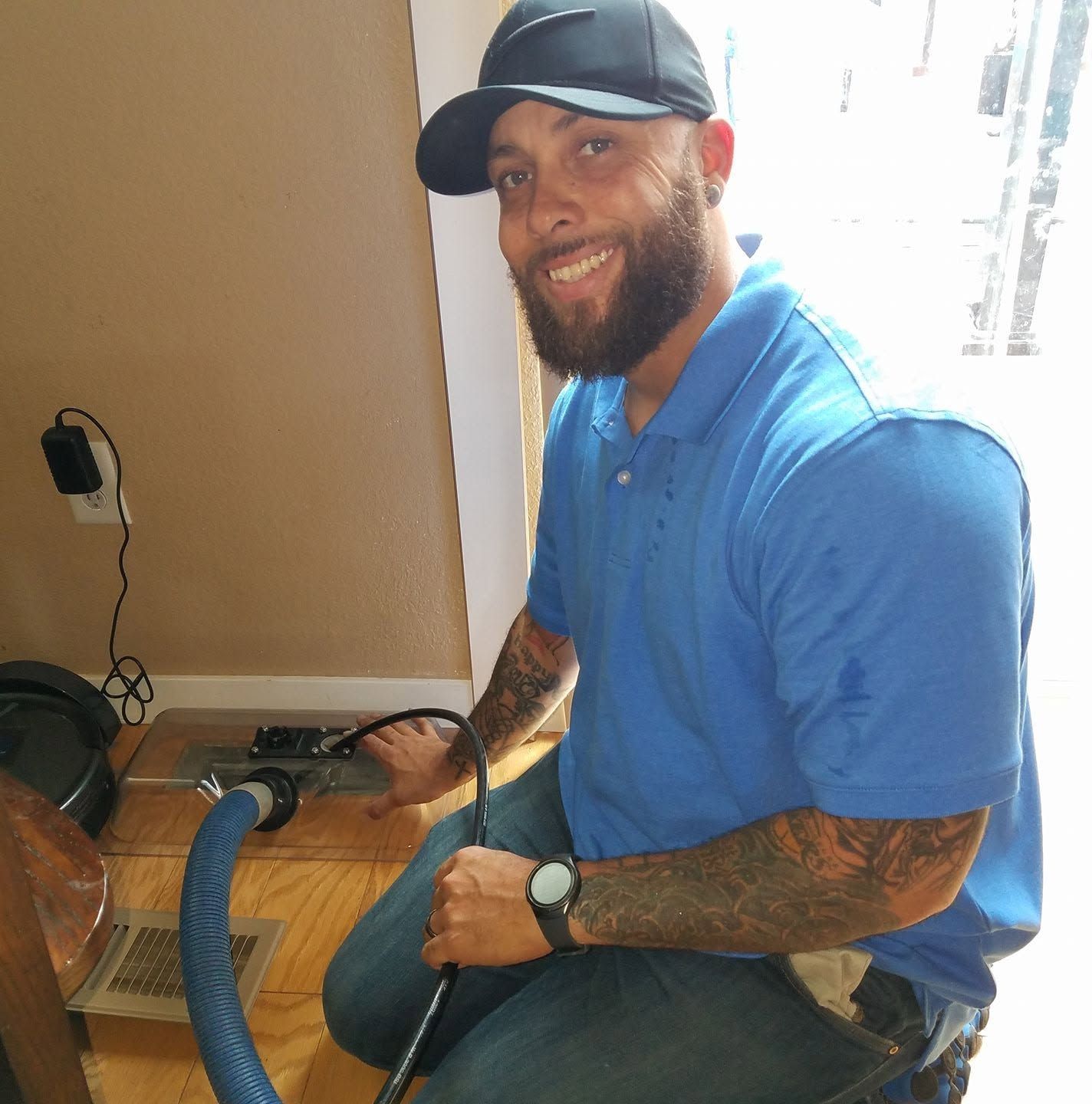 Man in blue polo and cap vacuuming beside a wall, smiling while kneeling on a hardwood floor.