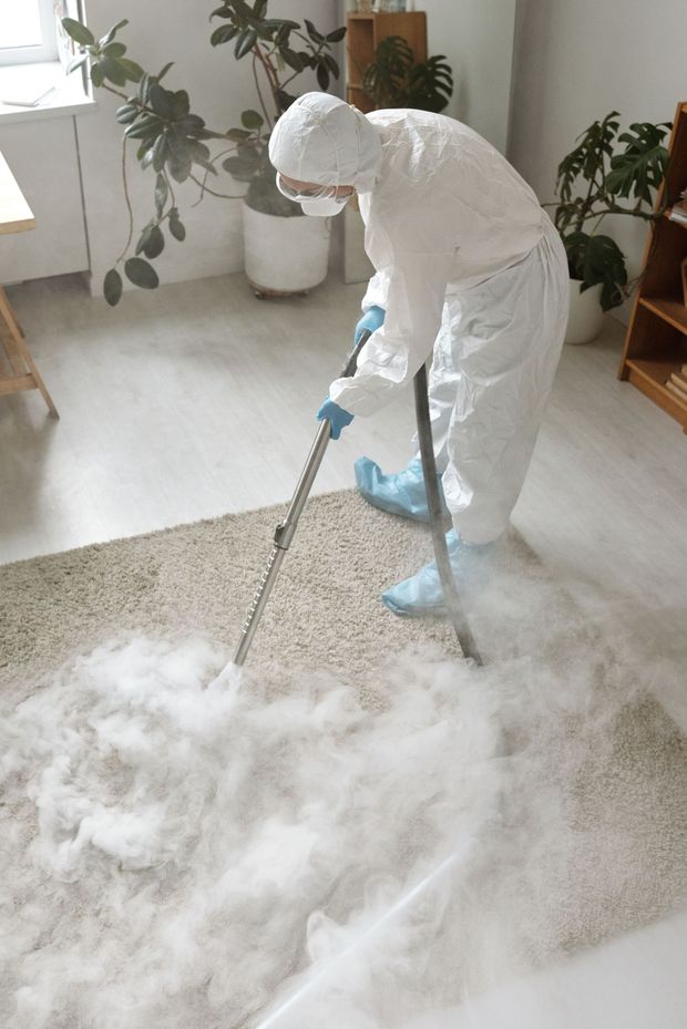 Person in white coveralls cleaning a carpet with a steam cleaner in a bright room