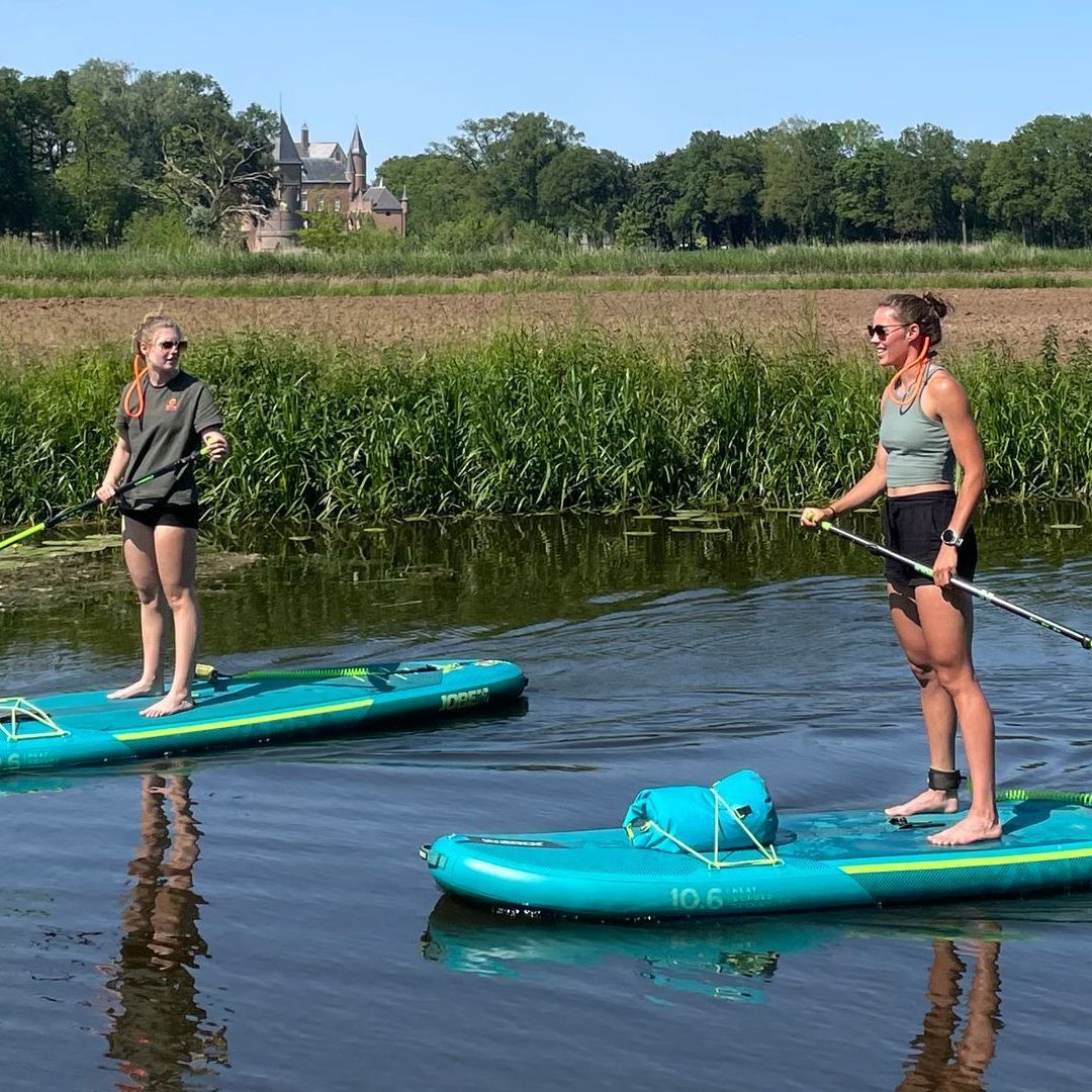 Twee mensen suppen op een rustige, smalle rivier, omzoomd door weelderige groene struiken en planten, onder een bewolkte hemel.