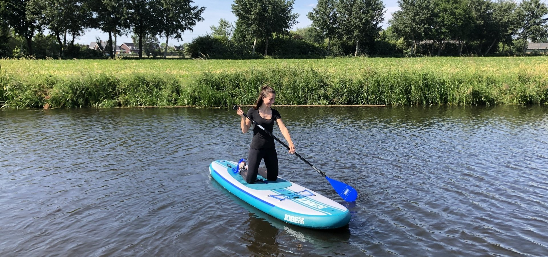 Een persoon knielt op een blauw paddleboard en gebruikt een peddel om zich voort te bewegen over een kalm, met gras omzoomd kanaal op een zonnige dag.