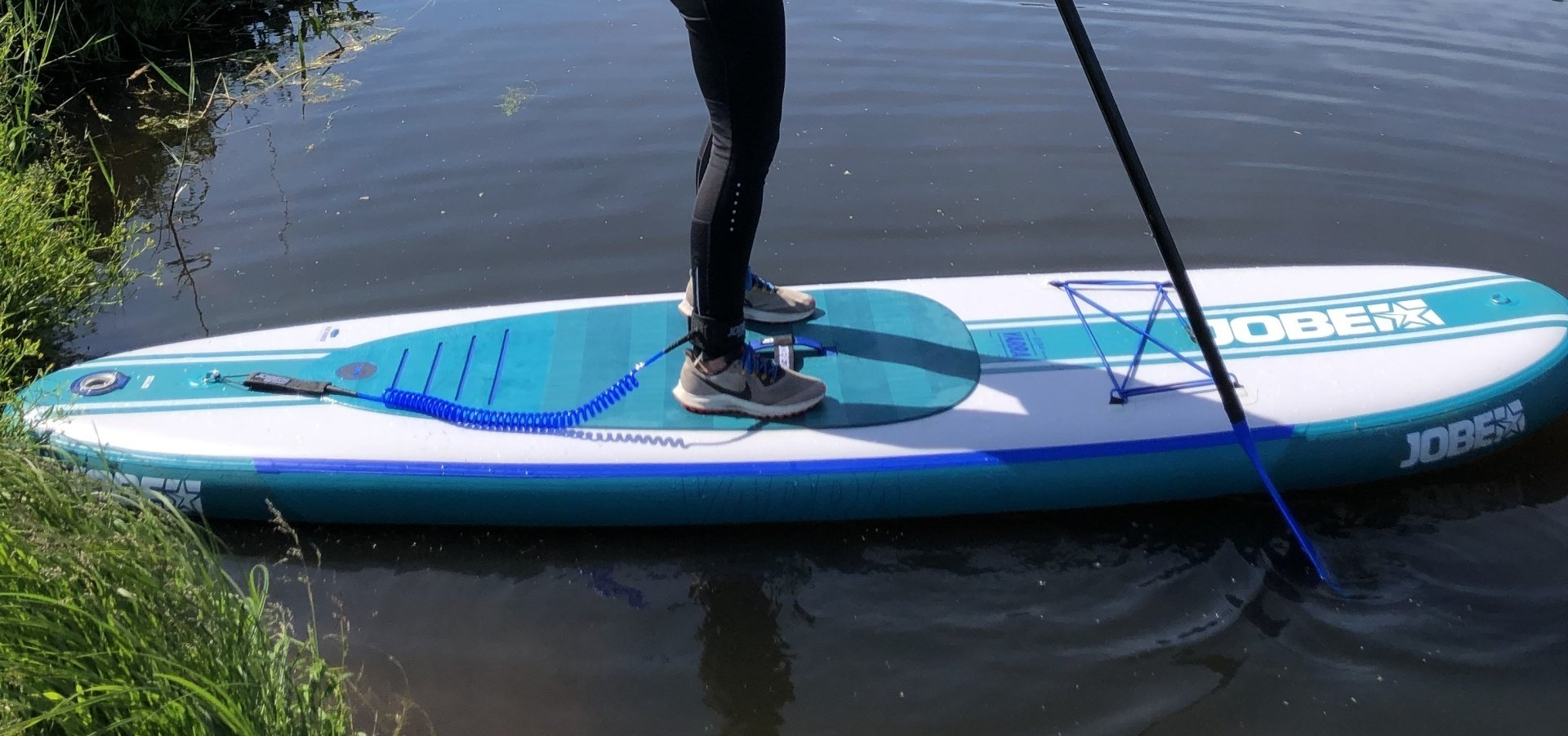 Een persoon staat op een blauw-wit Jobe stand-up paddleboard op rustig water vlakbij een grasrijke oever.