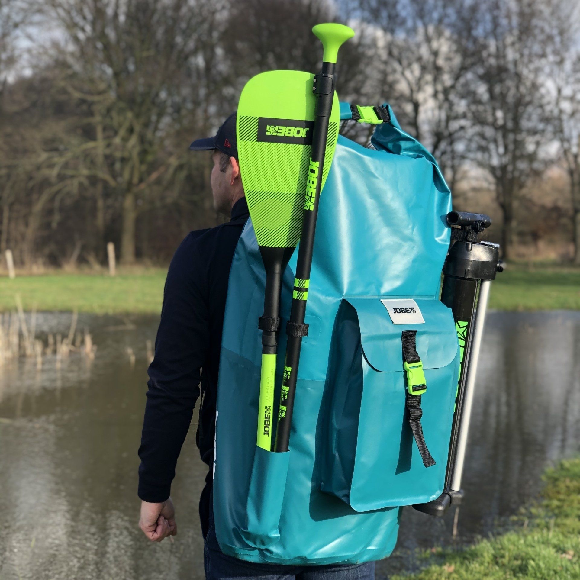 Een persoon draagt ​​een grote turquoise waterdichte rugzak met een daaraan bevestigde neon groene peddel en staat bij een vijver.