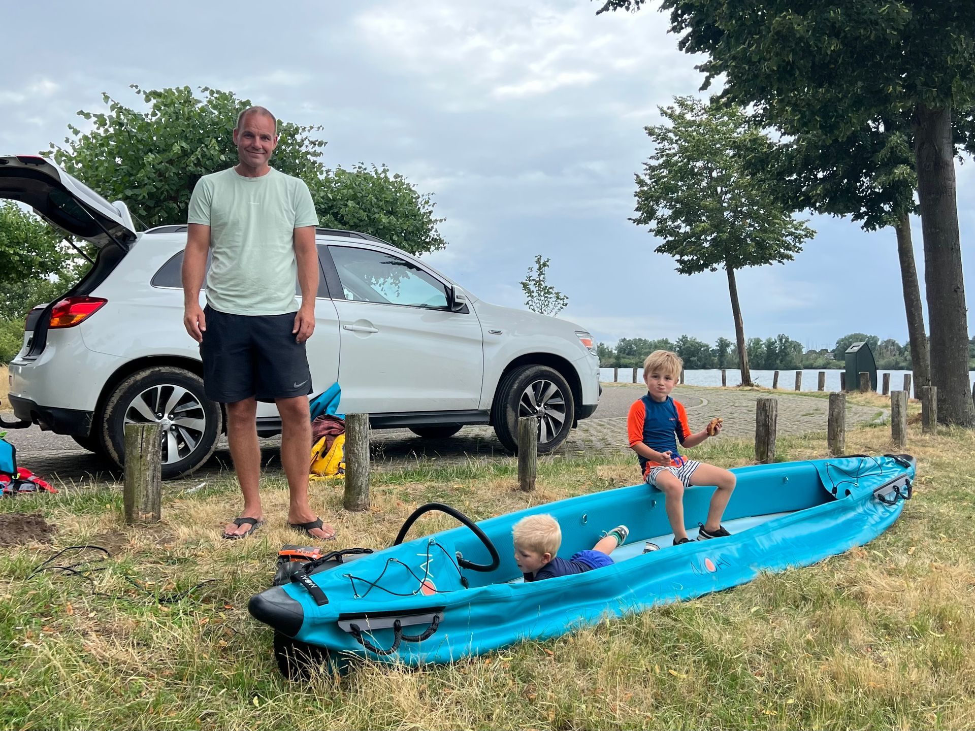 Een persoon staat naast een witte SUV aan de waterkant, terwijl twee kinderen in een felblauwe opblaaskajak zitten.