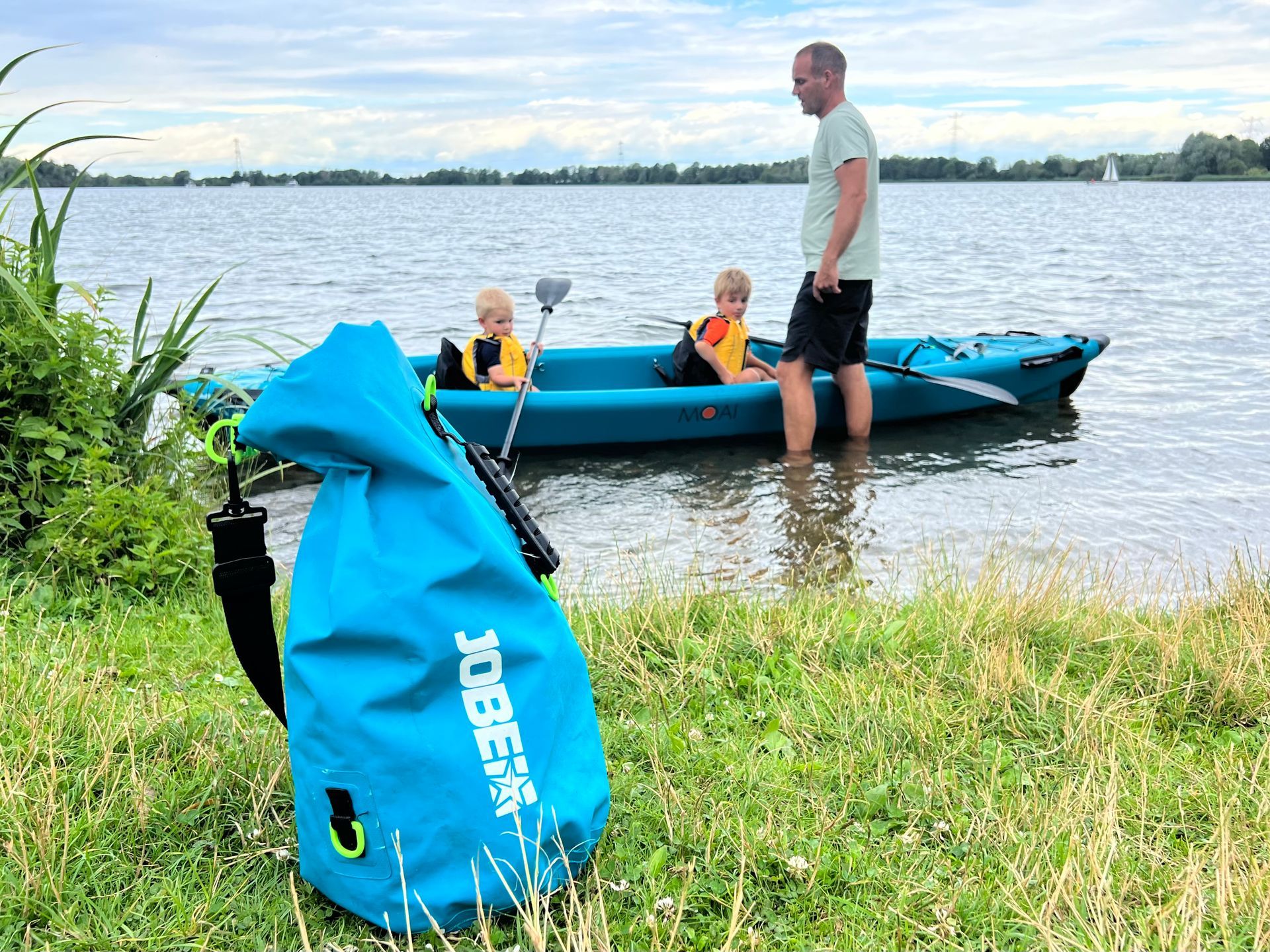 Een felblauwe, waterdichte tas staat op de grasrijke oever van een meer, met een man in een kajak en twee kinderen in de buurt.