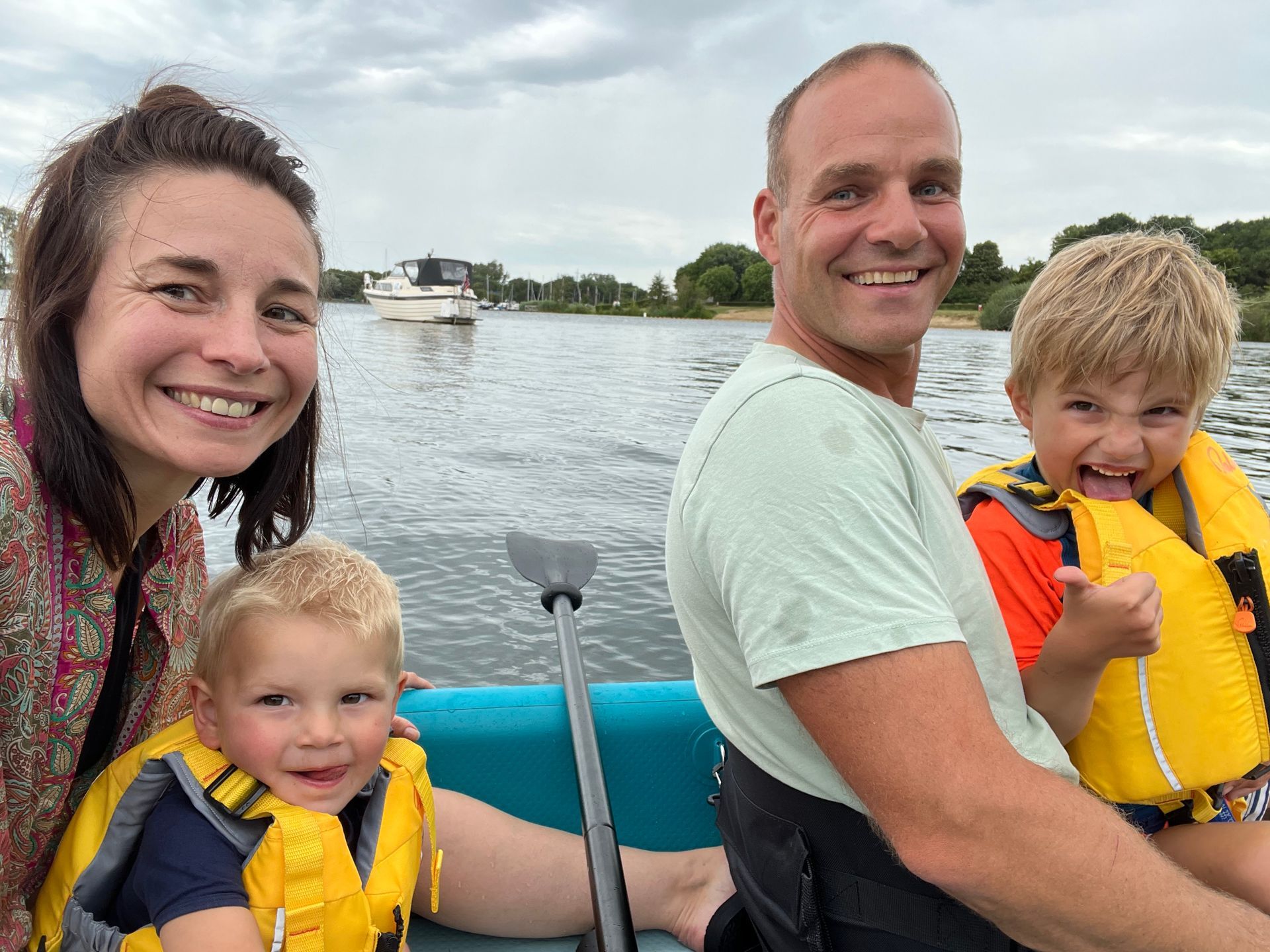 Een gezin van vier lacht terwijl ze in een blauwe boot op een meer peddelen, met een andere boot zichtbaar op de achtergrond.