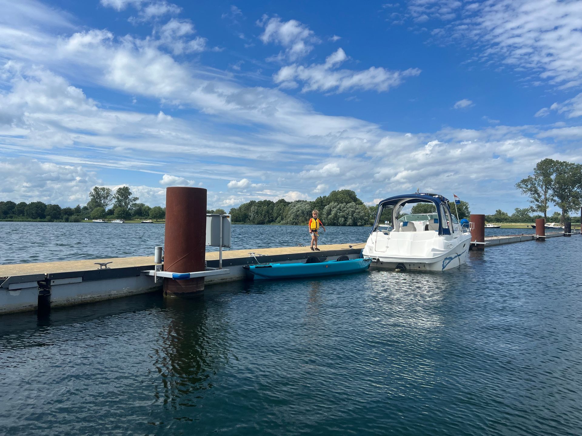 Op een zonnige dag staat een persoon met een reddingsvest op een houten steiger naast een blauwe kajak en een witte motorboot.