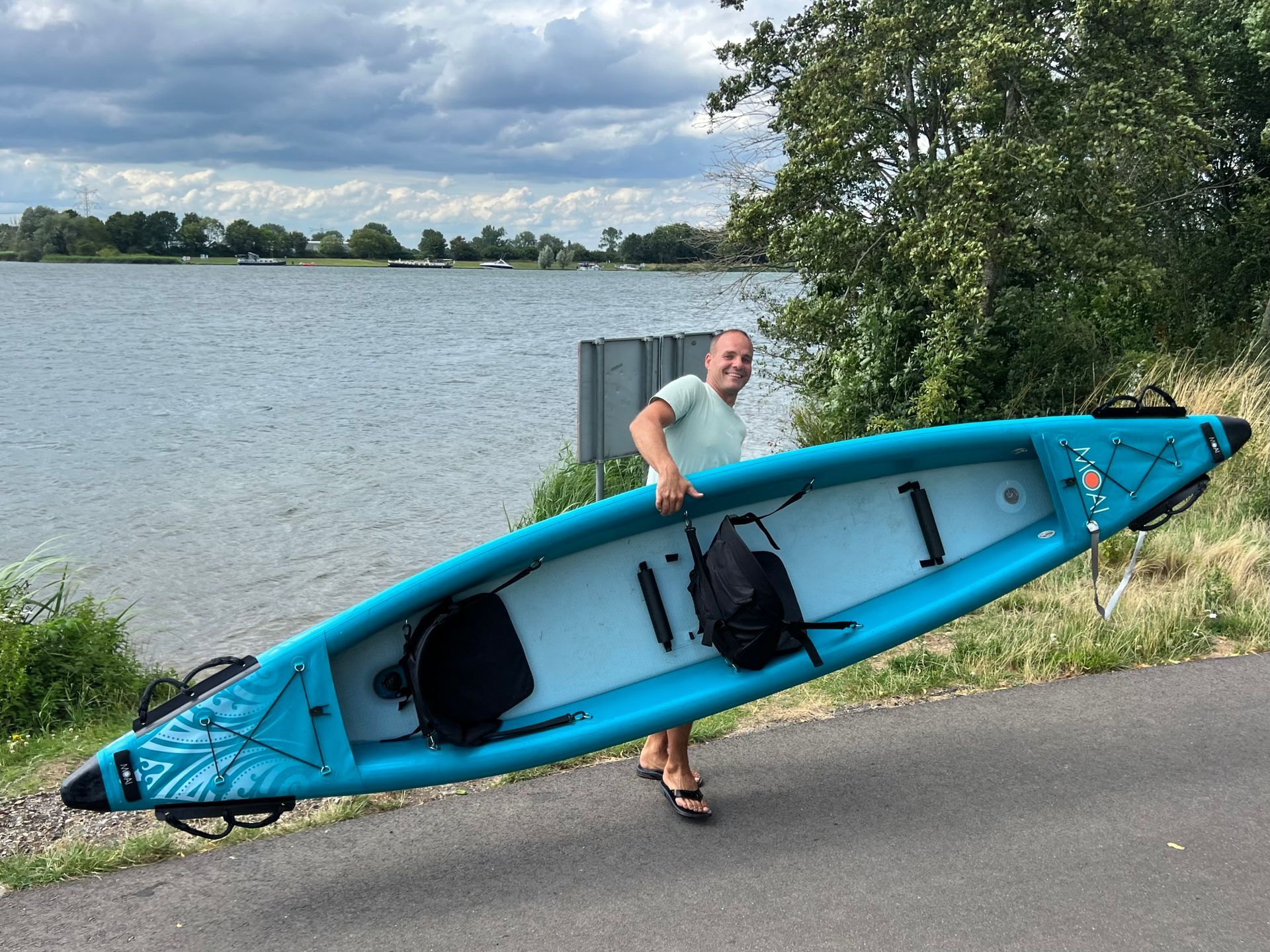 Een persoon lacht terwijl hij een turquoise opblaasbare kajak draagt ​​in de buitenlucht, vlakbij een groot wateroppervlak.