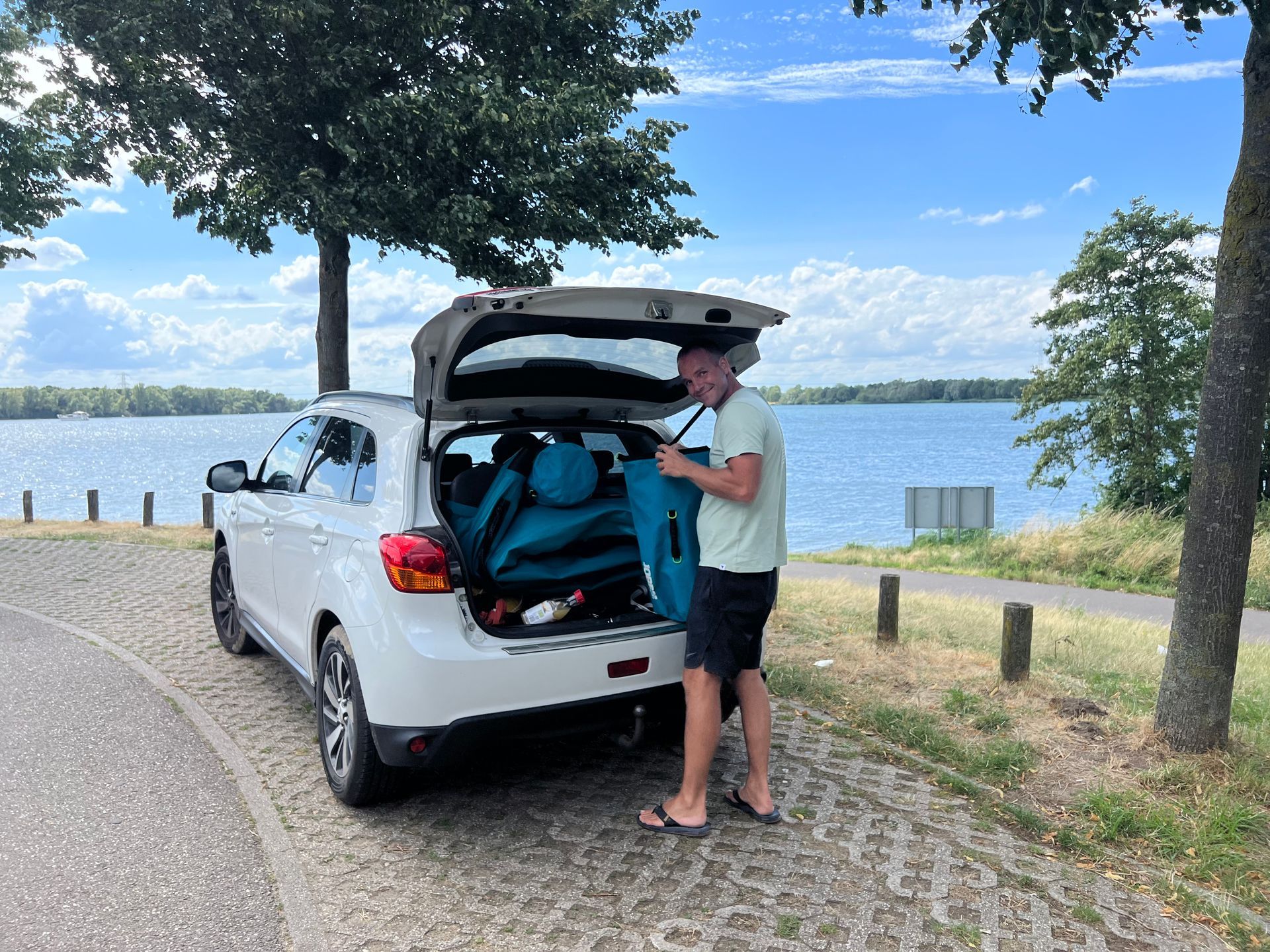 Een persoon in lichte kleding staat bij de open kofferbak van een witte auto die geparkeerd staat bij een meer, en pakt blauwe spullen in.
