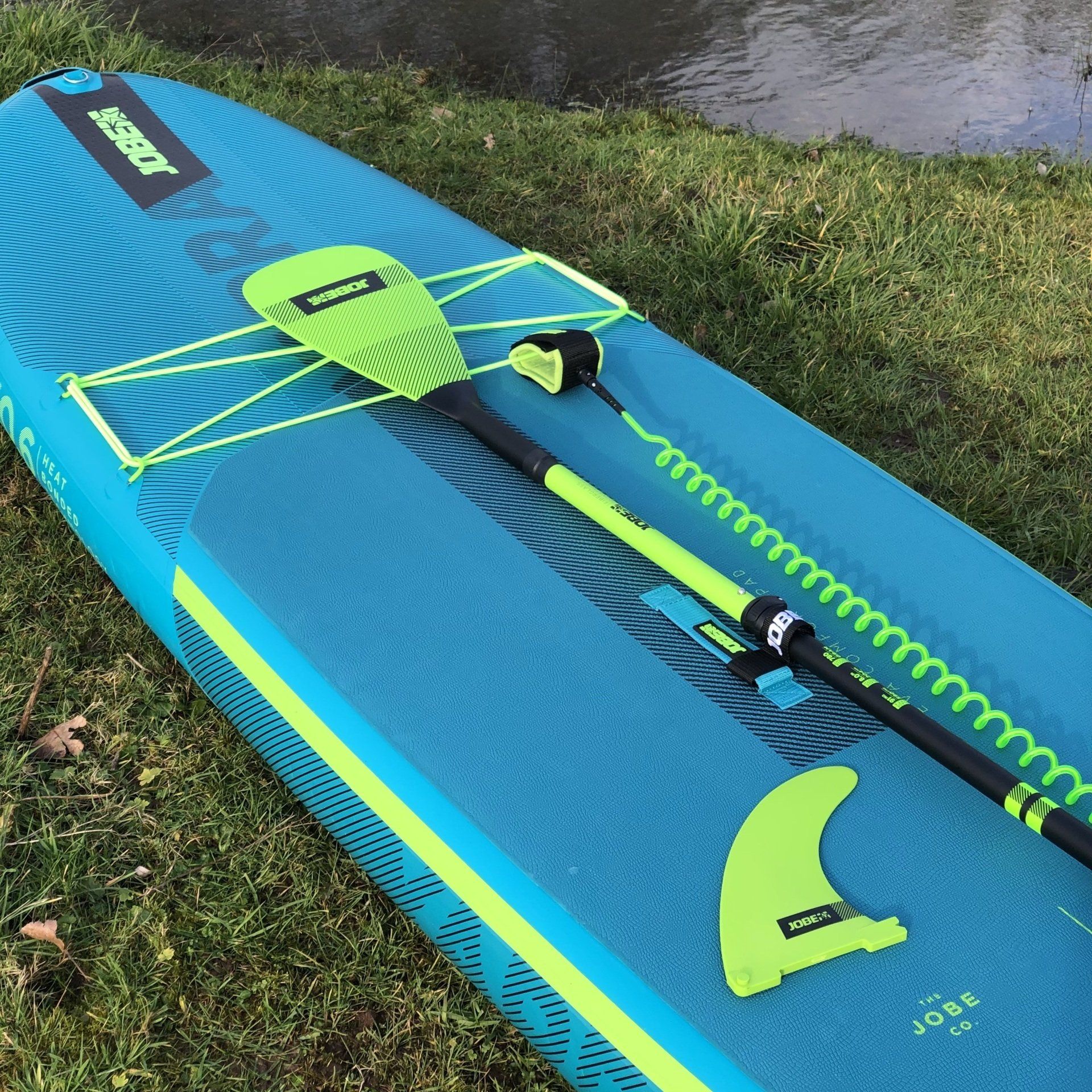 Een blauw Jobe stand-up paddleboard ligt op het gras, met een felgroene peddel, een opgerolde leash en een bijpassende vin erop.