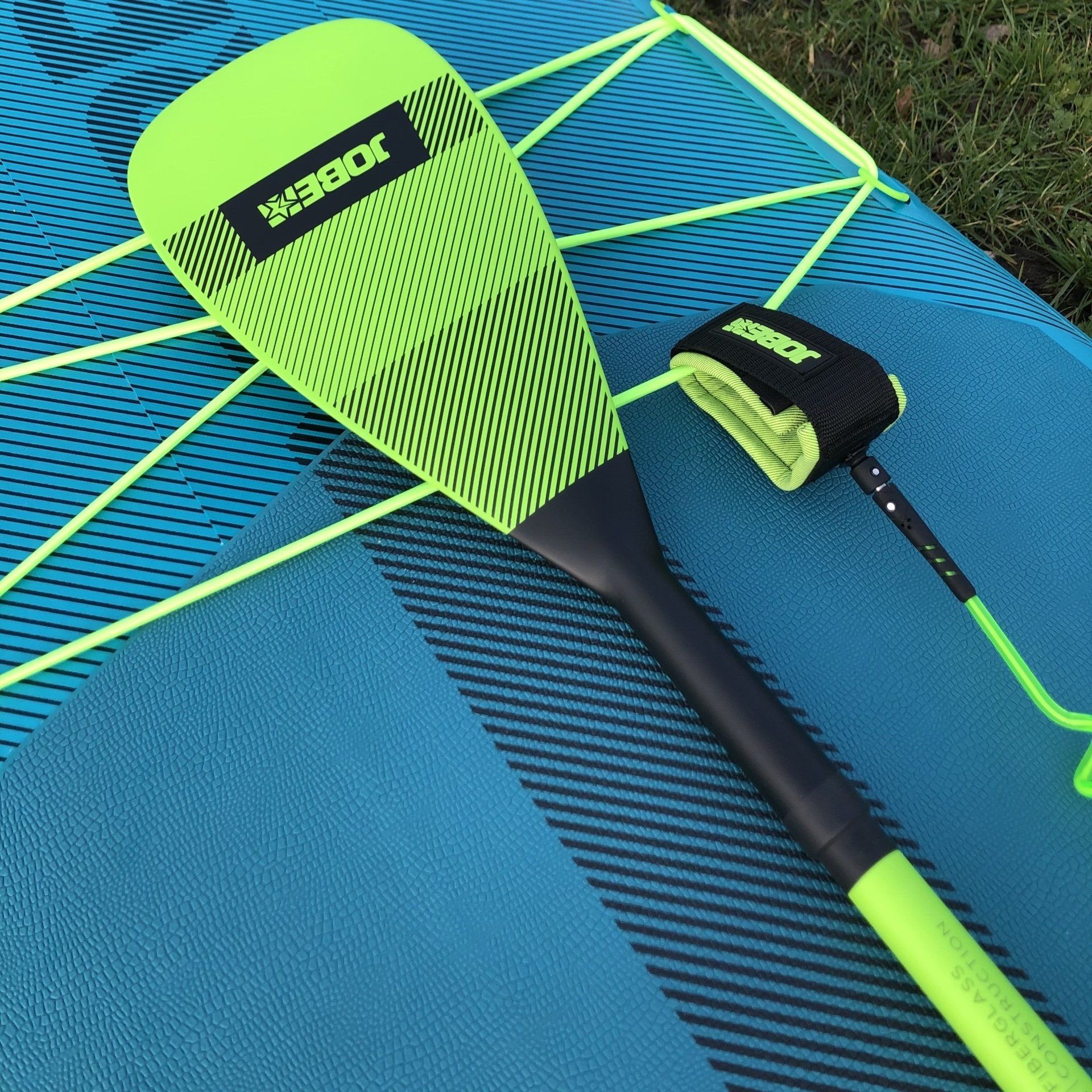 Een felgroene stand-up paddleboard peddel en een bijpassende enkelband liggen op een blauw, gestructureerd paddleboard.
