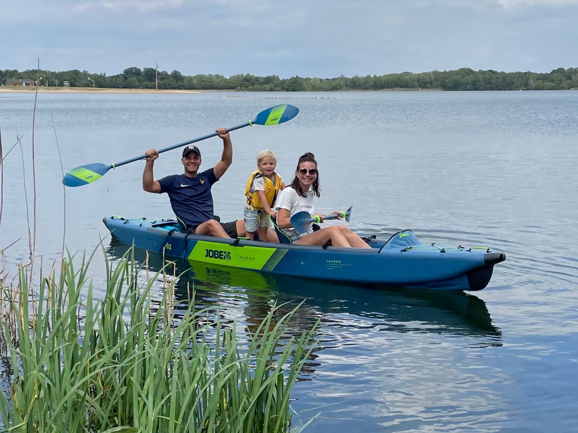 Drie mensen in een blauwe kajak op een meer, waarbij één van hen de peddel hoog in de lucht houdt tegen een bewolkte hemel.