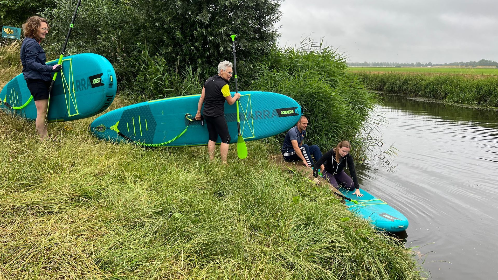 Op een bewolkte dag maken vier mensen turquoisekleurige stand-up paddleboards klaar langs een grasrijke rivieroever.