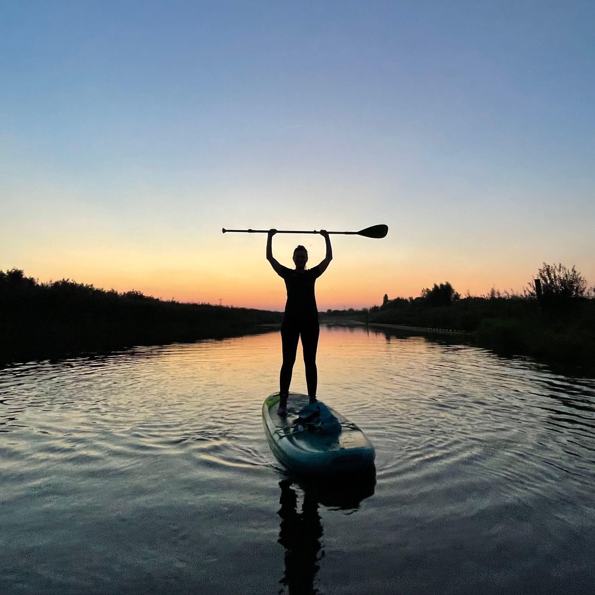 Een silhouet van een persoon die op een paddleboard op een rustige rivier staat en een peddel horizontaal boven het hoofd houdt.