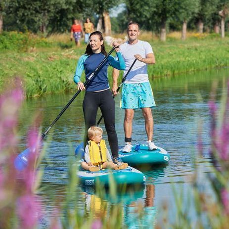 Twee mensen suppen op een kanaal, een van hen met een kind in een reddingsvest, op een zonnige dag met bomen op de achtergrond.
