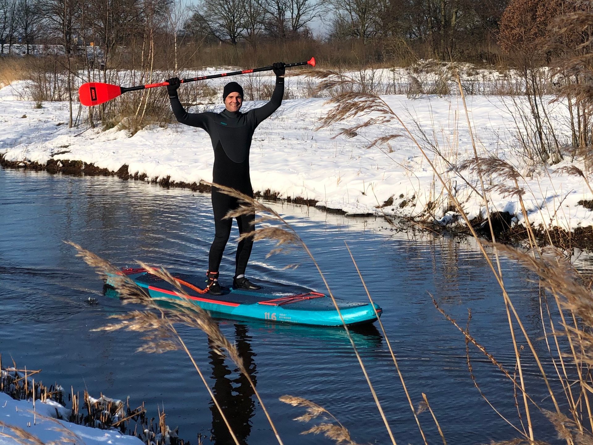 Een persoon in een zwart wetsuit staat op een blauw paddleboard in een besneeuwd landschap en houdt een peddel boven zijn hoofd.