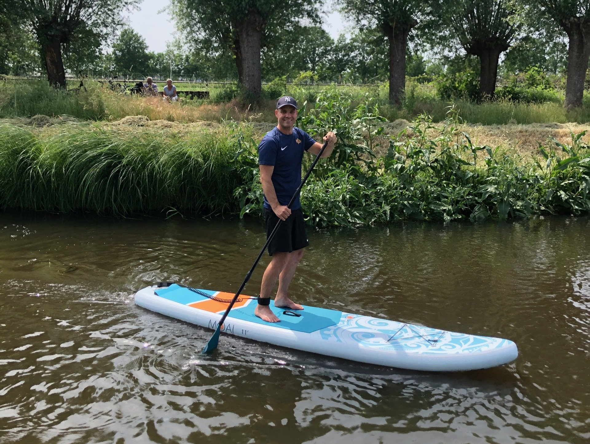 Een persoon die aan het suppen is op een rustig kanaal, peddelend, met weelderige groene bomen en grasrijke oevers op de achtergrond.