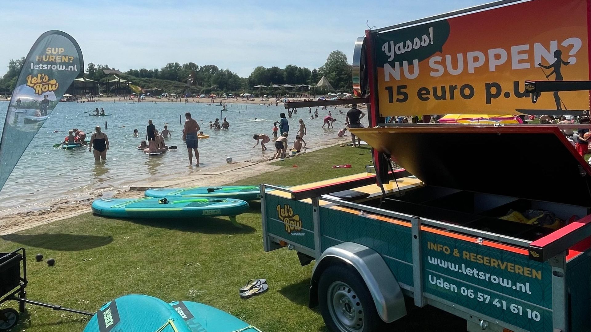 Een trailer met verhuur van stand-up paddleboards op een zonnig strand aan een meer, waar mensen zwemmen en ontspannen in het water.