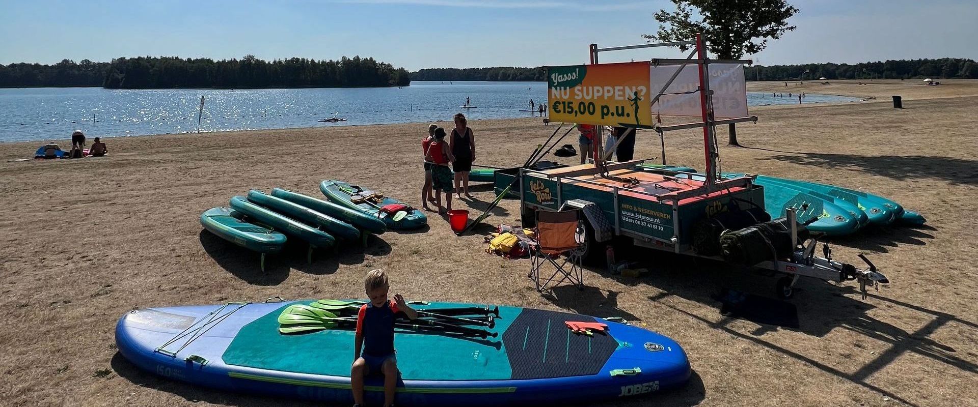 Een strandscène met een aanhanger voor de verhuur van stand-up paddleboards, met diverse boards die op het zand bij een meer liggen.