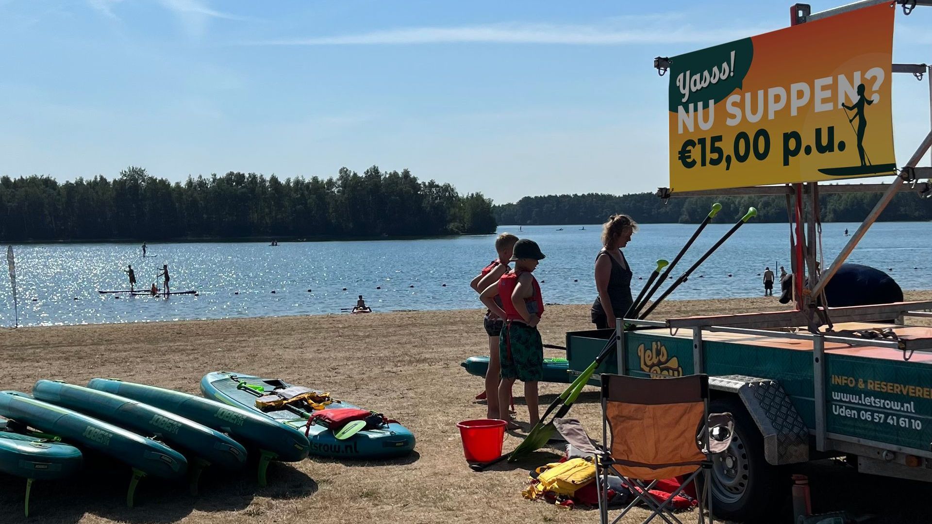 Een verhuurstation voor stand-up paddleboards aan een zandstrand aan een meer, met een bord waarop sessies voor €15 per stuk worden aangeboden.