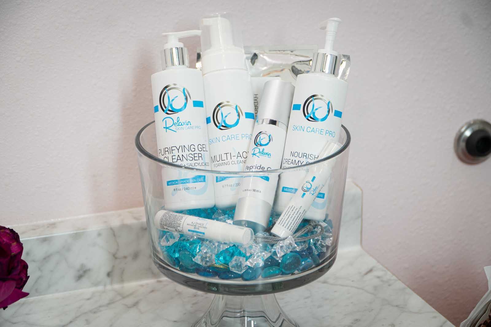 Skincare products in a glass bowl filled with blue decorative beads, on a marble countertop.