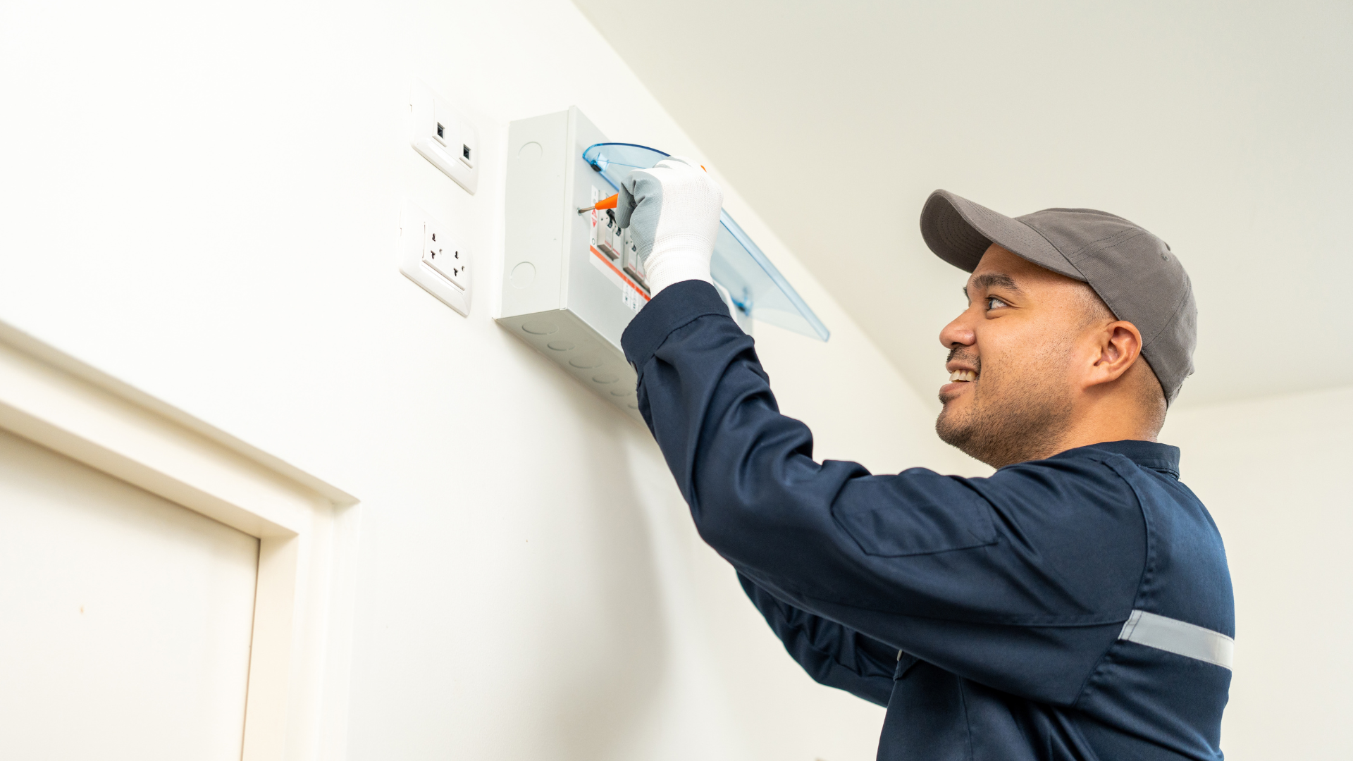 Man installing a smoke detector on a white wall, smiling and wearing a cap and dark shirt