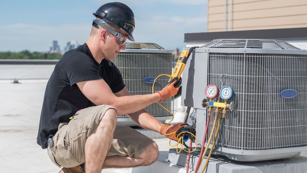 Technician kneels on rooftop inspecting HVAC unit with gauges and tools connected to pipes