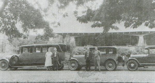 Vintage cars parked on a road with people standing beside them. A house is in the background.