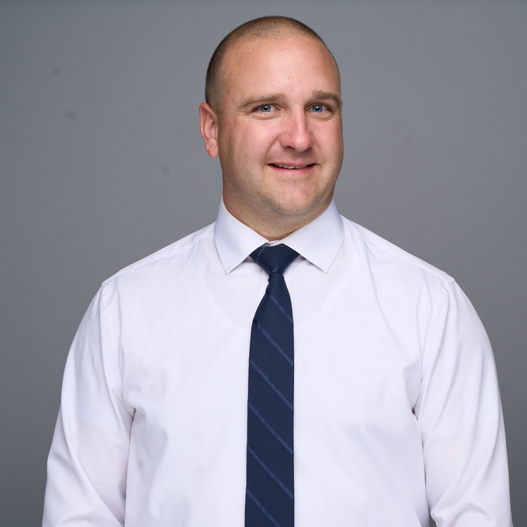 Man in a white shirt and navy tie, smiling against a gray background