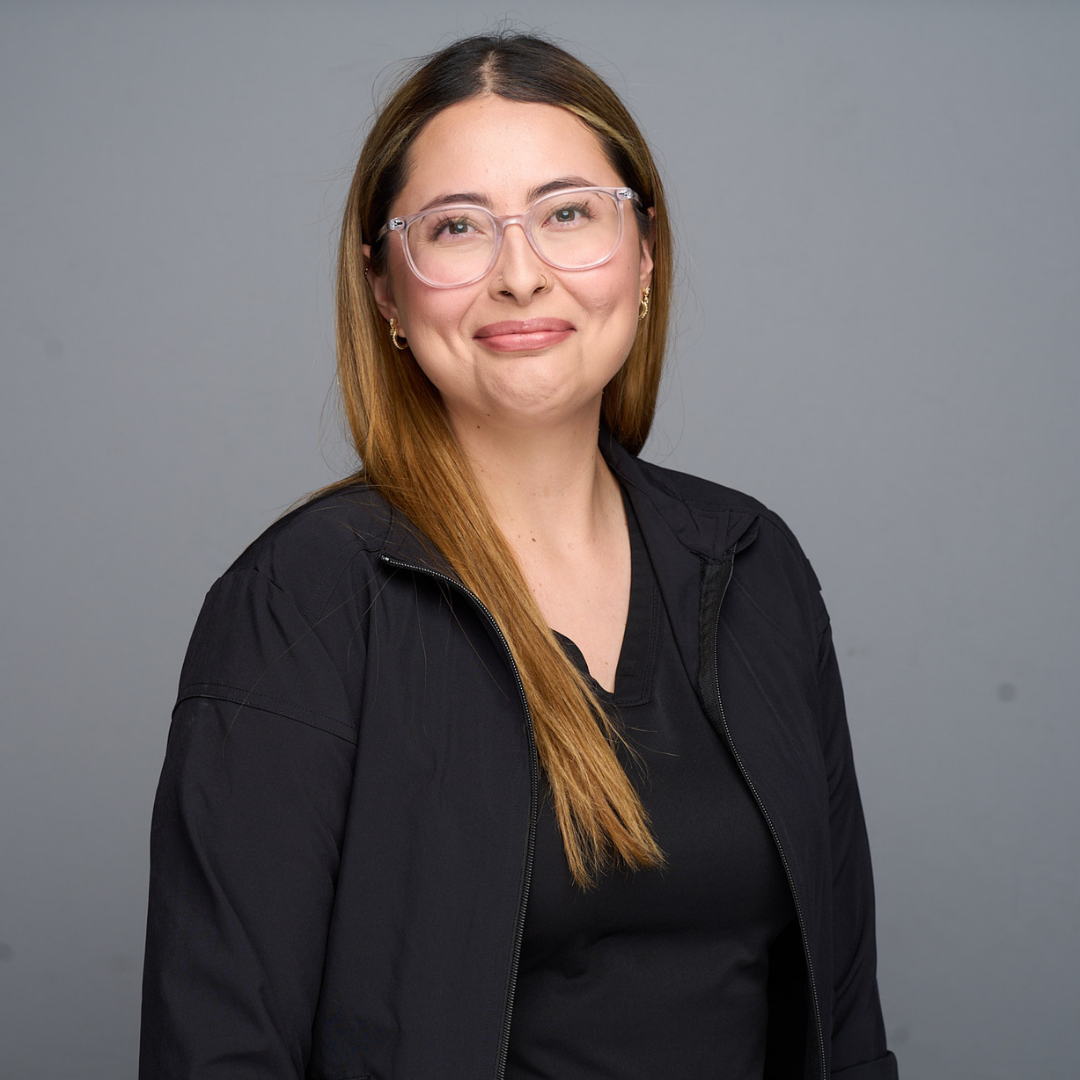 Smiling person with glasses and long hair, wearing a black jacket, standing against a gray background