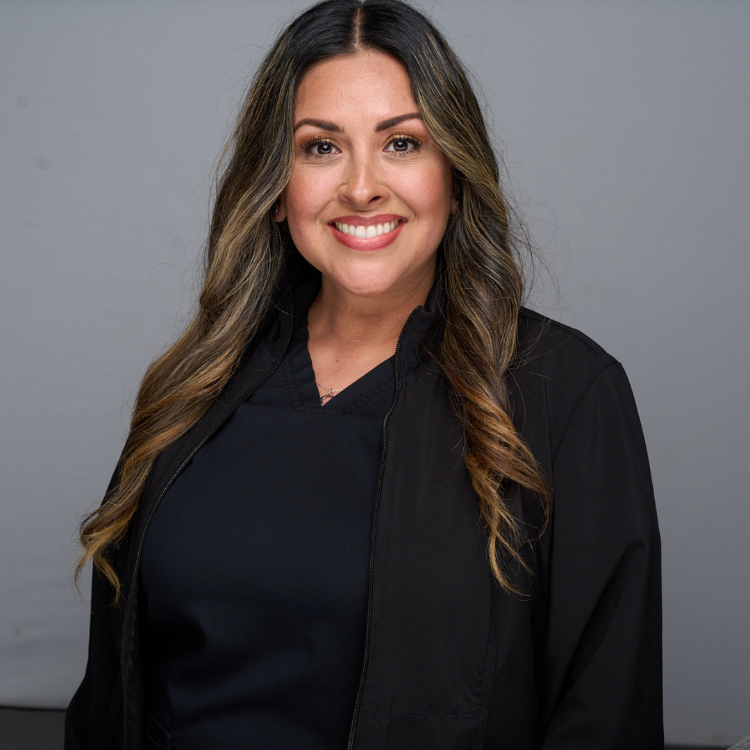 Smiling woman with long dark hair wearing a black top and jacket against a gray background
