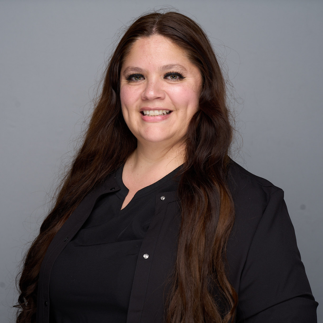 Smiling person with long brown hair in a black top and blazer against a gray background