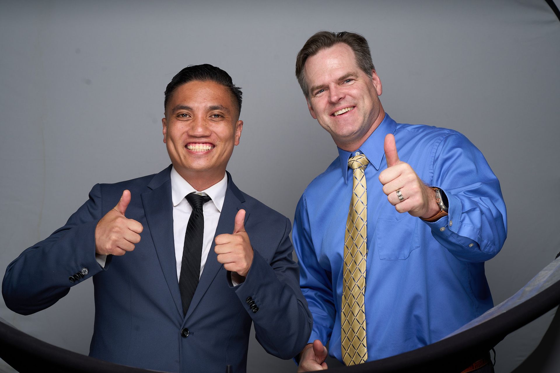 Two men in business attire smiling and giving thumbs up against a plain gray backdrop