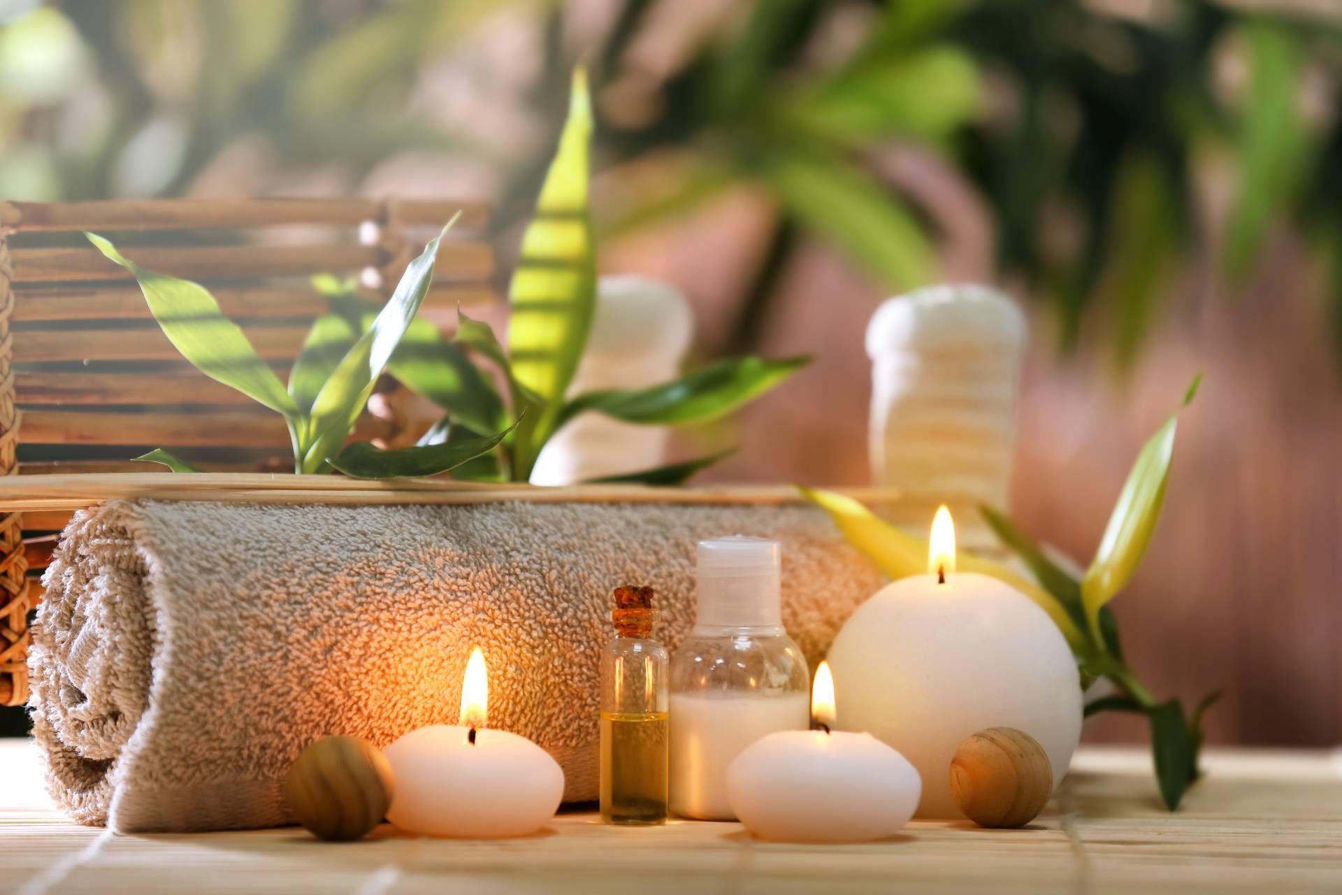 Spa scene with rolled towel, lit candles, and small bottles beside green leaves on a wooden surface