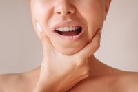 Close-up of a person touching their jaw and cheek, mouth open against a plain background