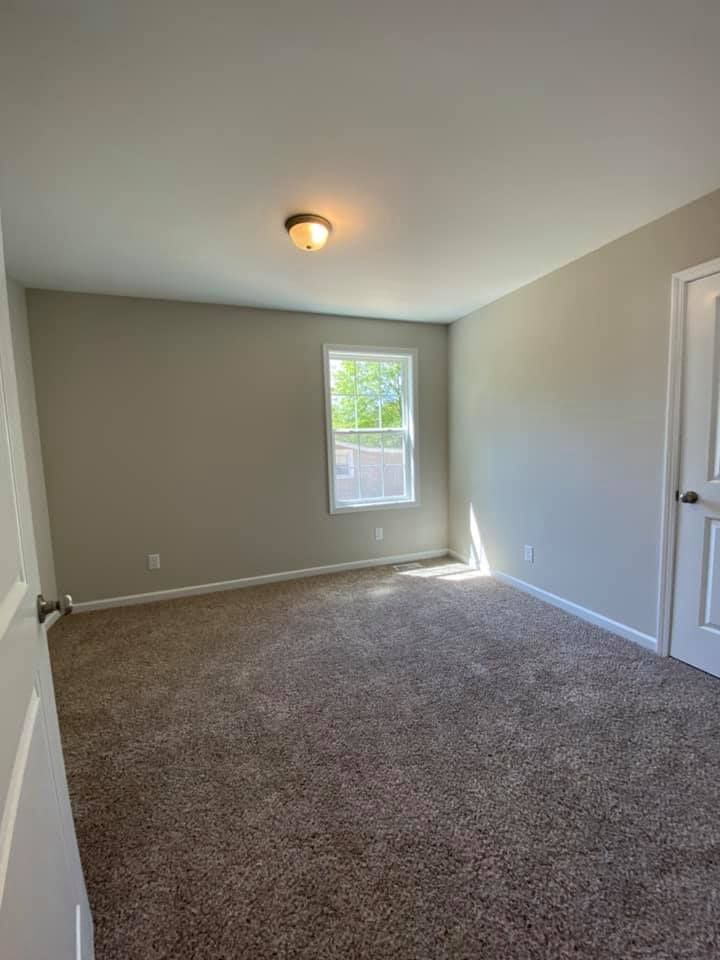 Empty beige bedroom with carpet, white trim, single window, and ceiling light.