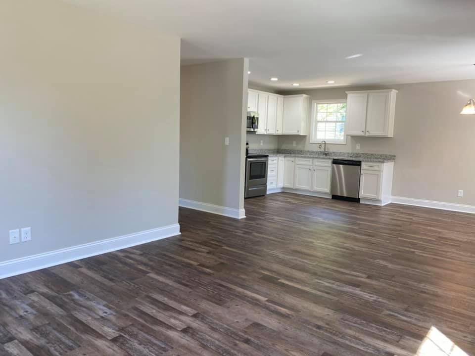 Empty open-plan living area with wood floors, white walls, and a small kitchen in the back.