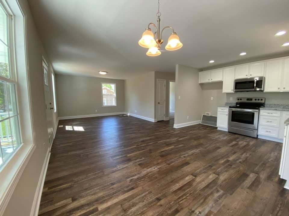 Open-concept living room with dark wood floors, white walls, and a small kitchen area with white cabinets.