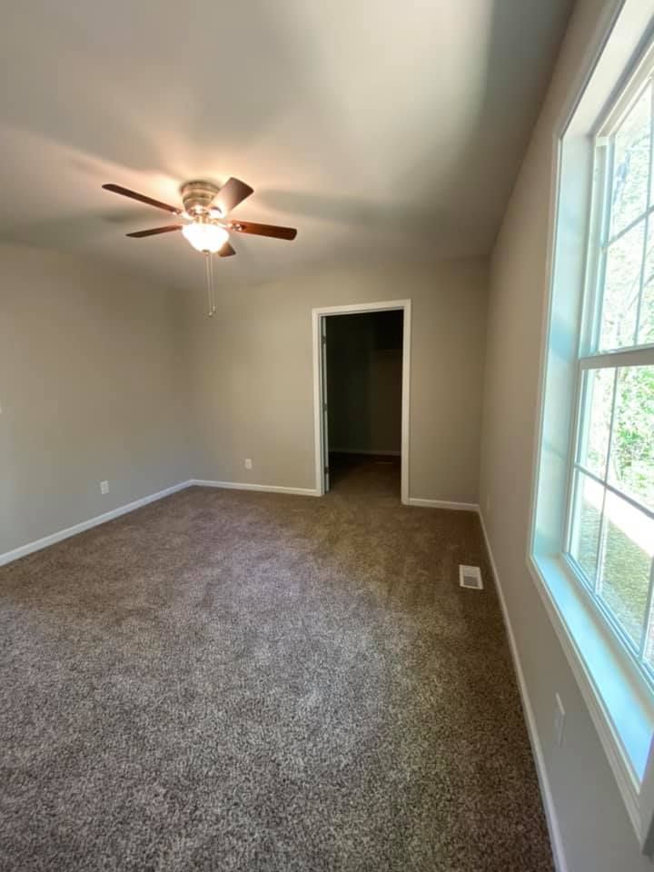 Empty carpeted bedroom with ceiling fan, beige walls, and a large window letting in daylight