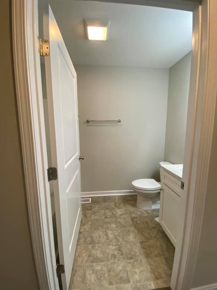 Small bathroom with toilet, vanity, towel rack, and beige tile floor viewed from the doorway.