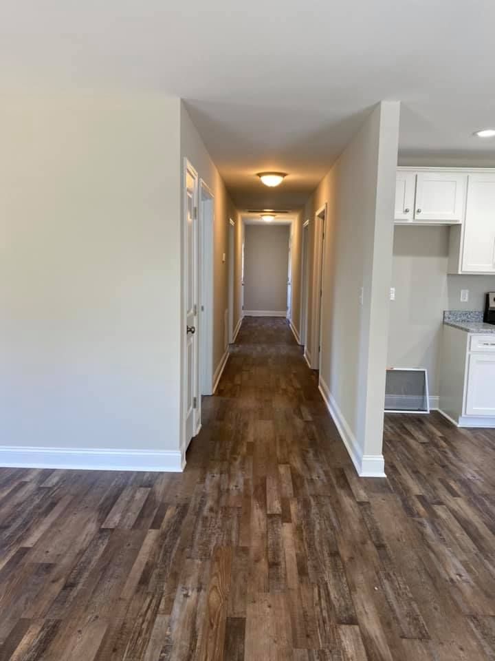 Empty hallway with wood flooring, white walls, and a view into a bright kitchen area.