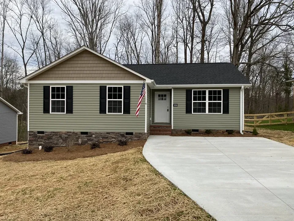 Single-story beige house with white trim, front porch, American flag, and a wide concrete driveway