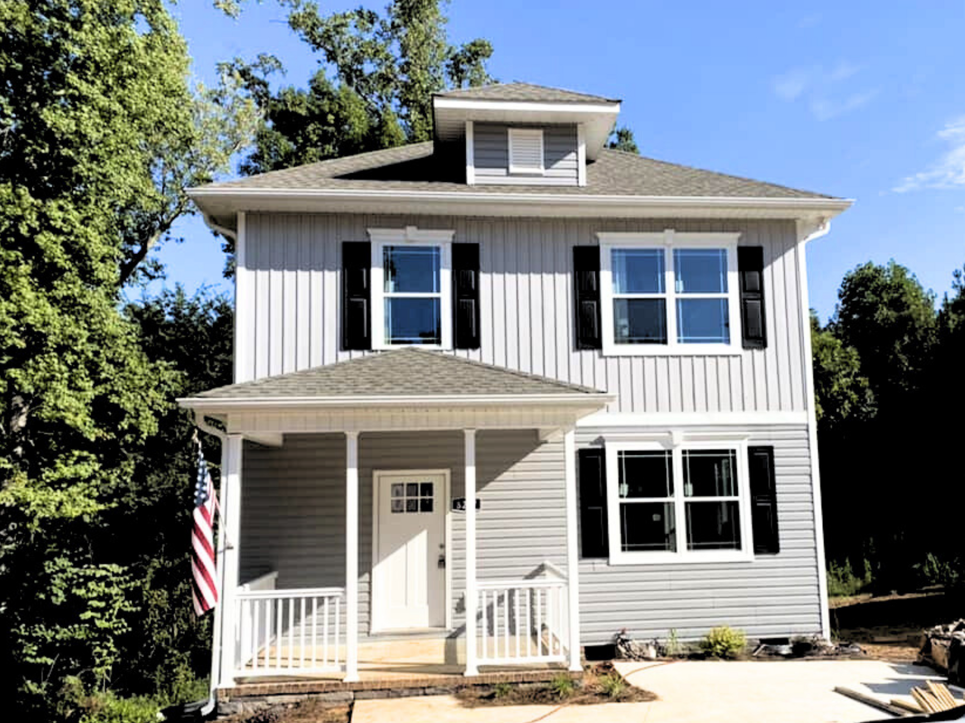 Two-story white house with black shutters, front porch, and American flag in bright daylight