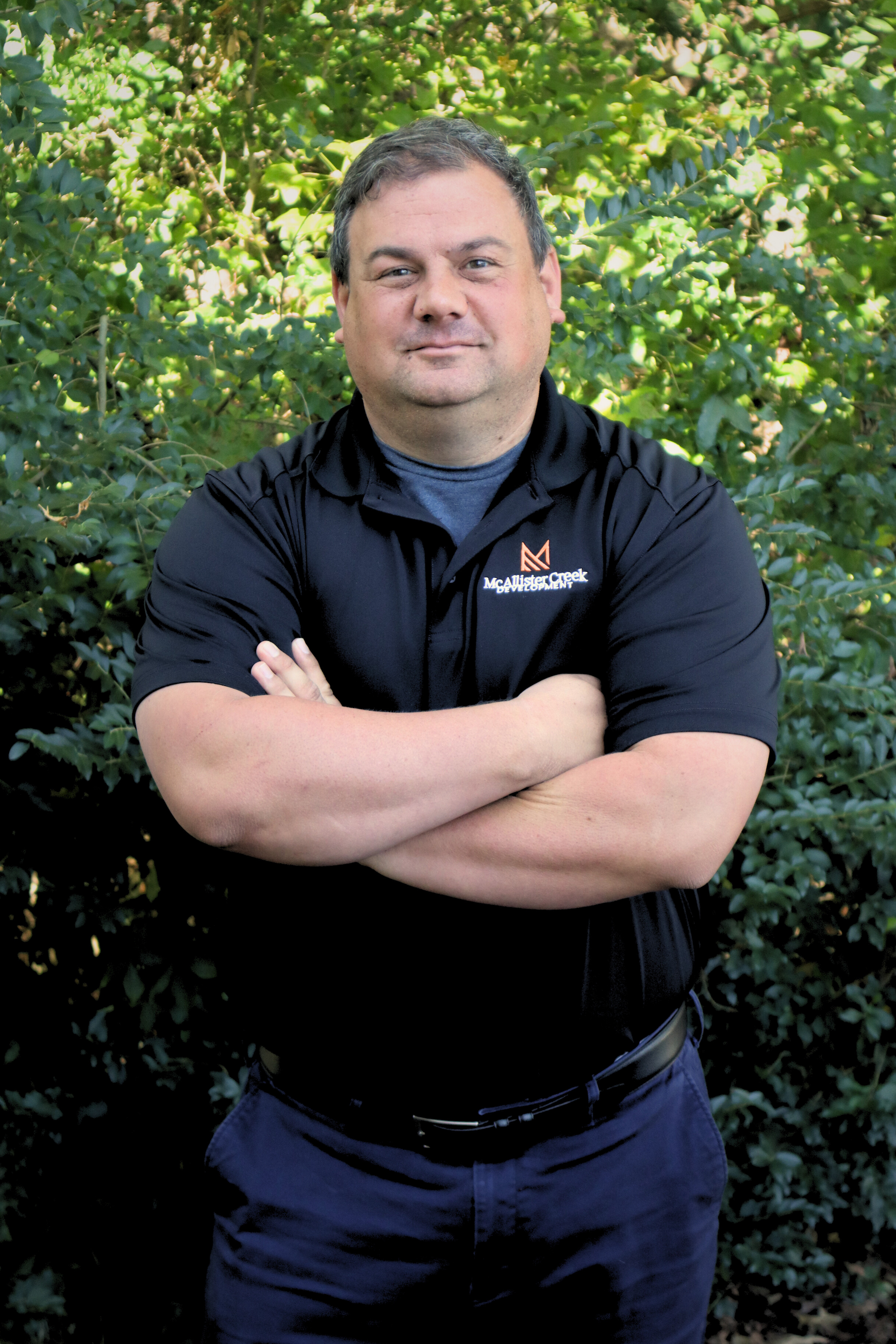 Man standing with arms crossed in a black polo shirt against leafy green bushes.