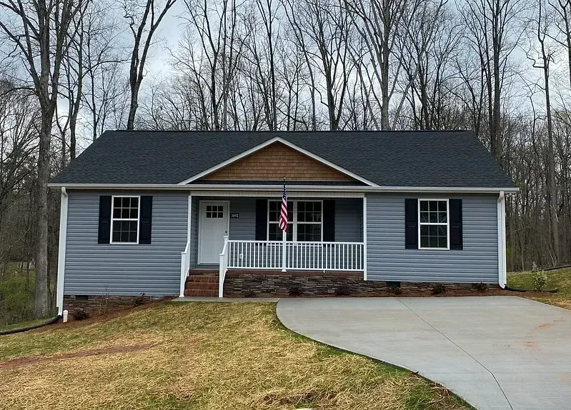 Single-story gray house with white trim, front porch, and a curved driveway in a wooded yard.