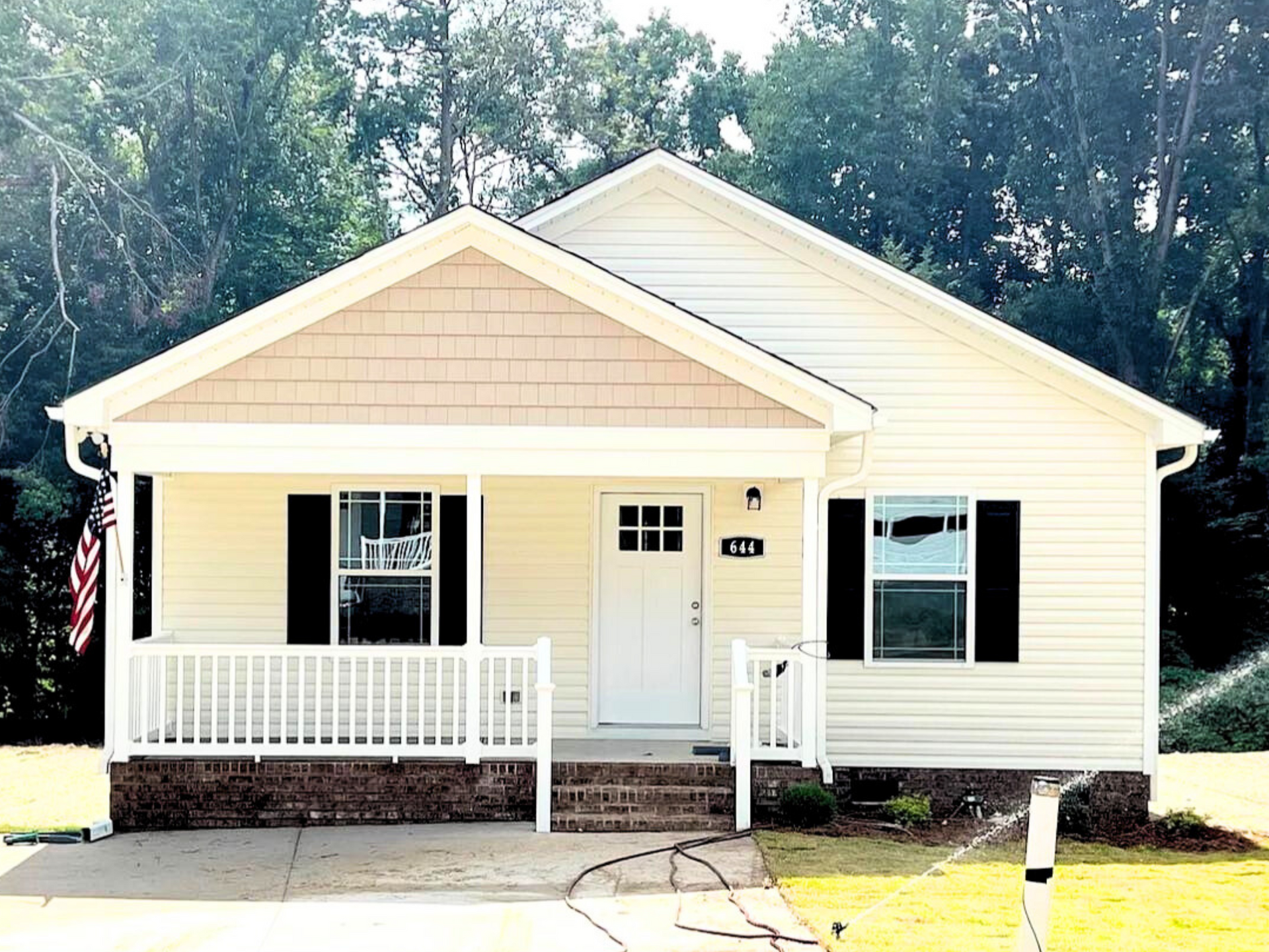 Small yellow house with white trim, front porch, and black shutters in a wooded yard
