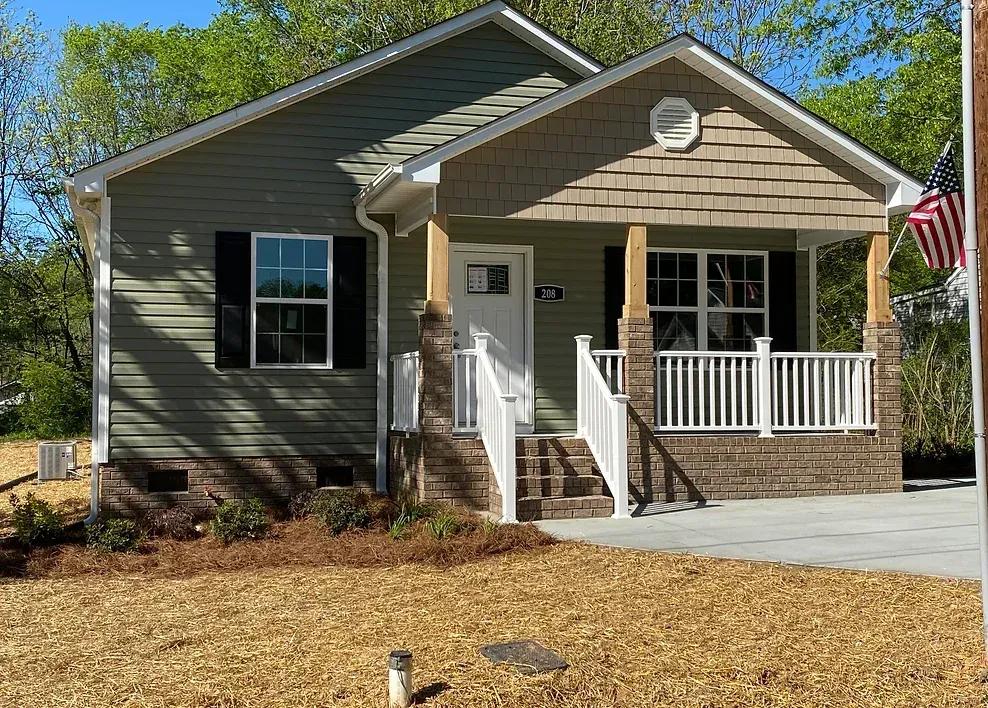 Single-story gray house with a front porch, white railings, steps, and an American flag in the yard