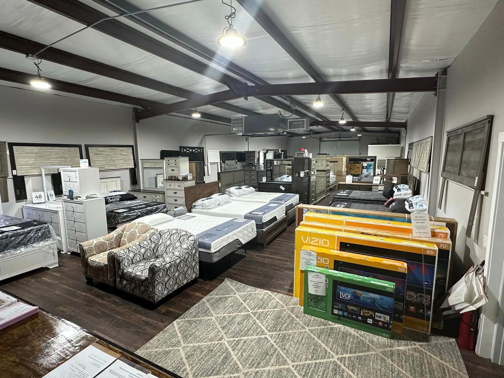 Furniture showroom with multiple beds, mattresses, two patterned armchairs, and stacks of packaged electronics on a rug.