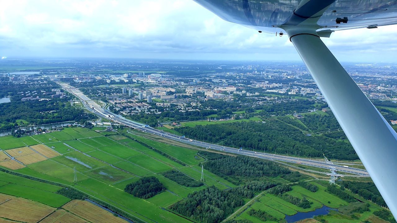 Luchtfoto van groene velden en een stadsgezicht, gezien vanuit een vliegtuigvleugel boven een kustgebied.